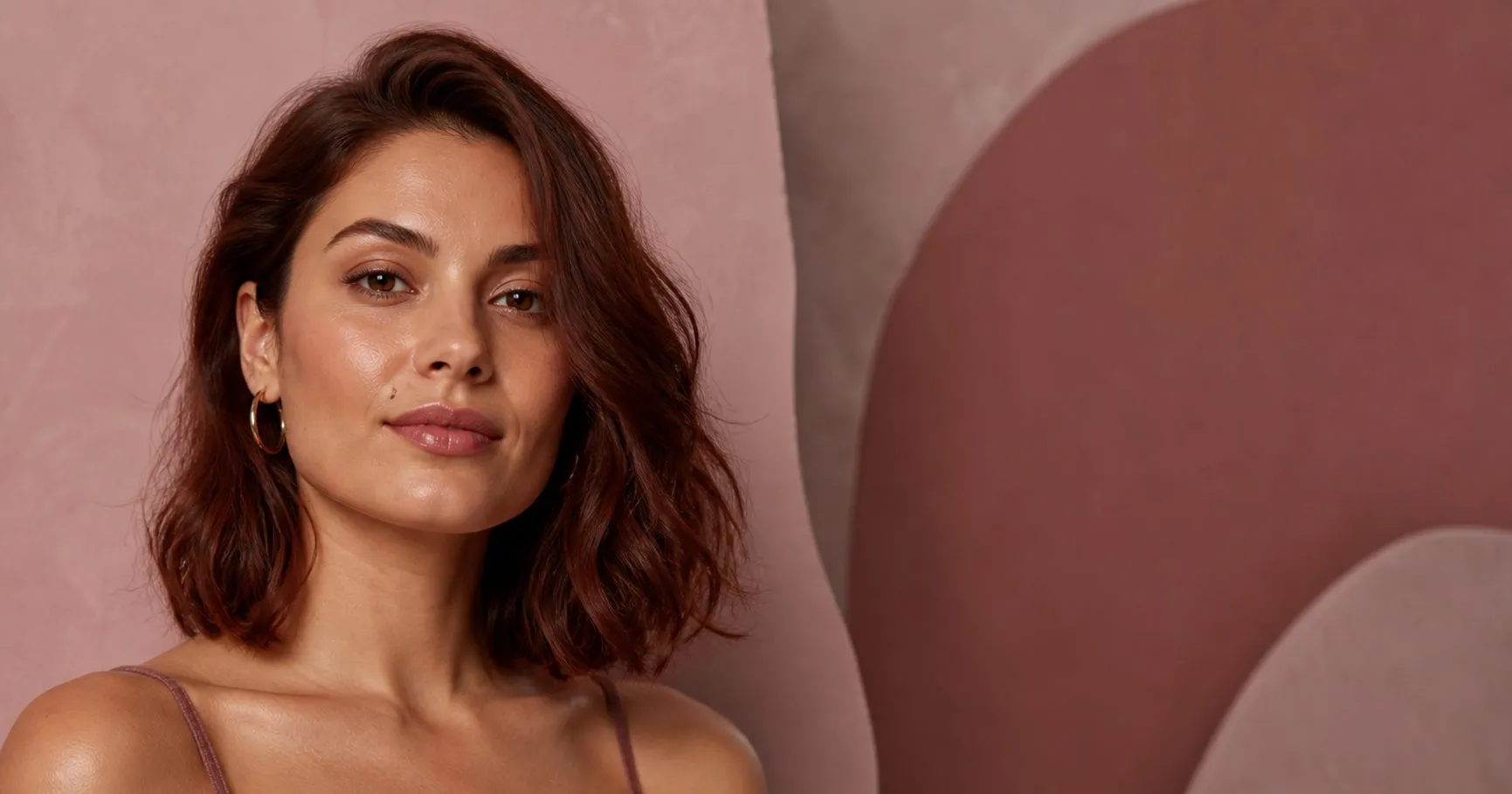 Young woman with a square face wearing a dark-auburn textured collarbone lob against a sculptural blush and mauve backdrop