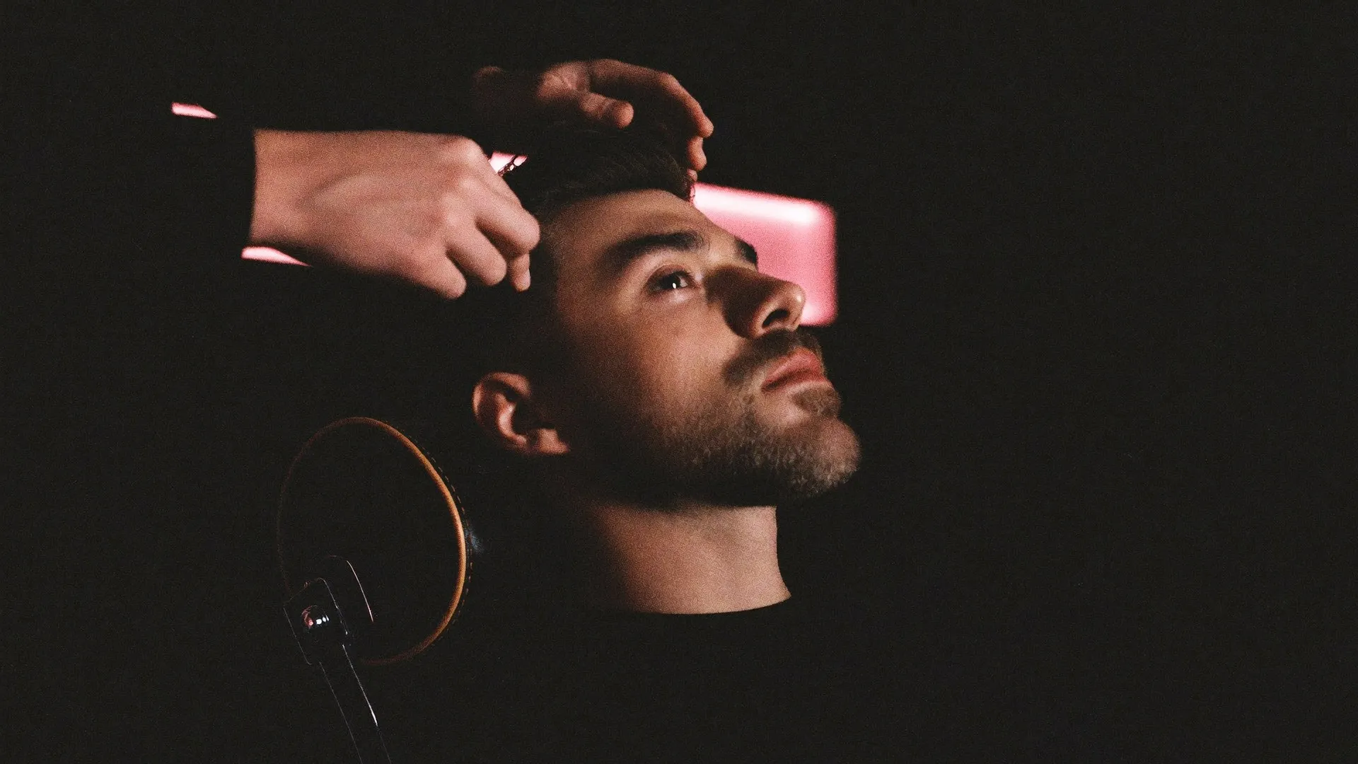 Man in a barbershop chair with a textured crop being adjusted by a barber under low pink light